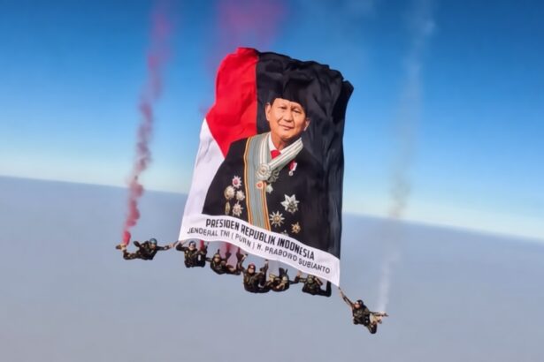 Skydivers hold up a large banner with a portrait of a male official in ceremonial dress, red-and-black flag colors, and Indonesian presidential text.