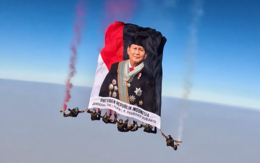 Skydivers hold up a large banner with a portrait of a male official in ceremonial dress, red-and-black flag colors, and Indonesian presidential text.