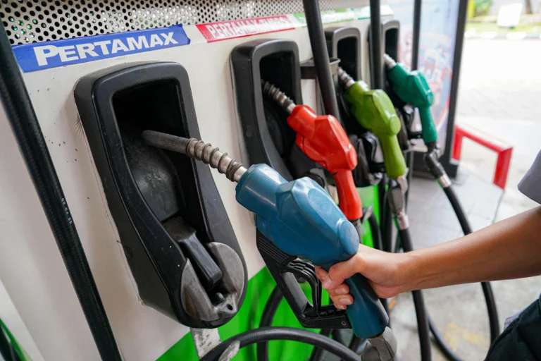 Person refueling a vehicle with a blue gasoline nozzle at a pump; several colored nozzles are visible in the background.