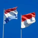 Two Dutch flags fluttering on flagpoles against a clear blue sky.