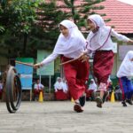 Two schoolgirls in white hijabs and red uniforms skip with sticks while a tire rolls nearby during a school event.