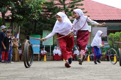 Two schoolgirls in white hijabs and red uniforms skip with sticks while a tire rolls nearby during a school event.