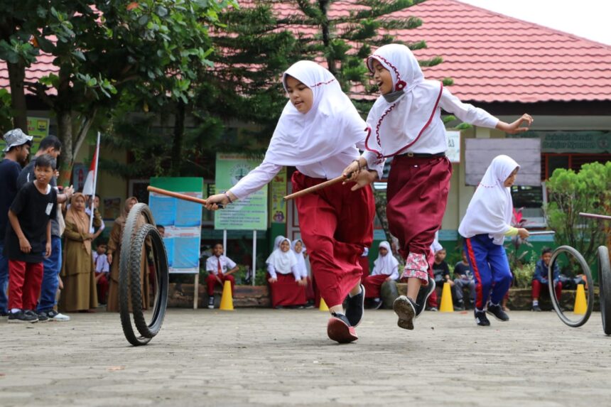 Two schoolgirls in white hijabs and red uniforms skip with sticks while a tire rolls nearby during a school event.