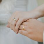 Close-up of a couple's hands with wedding bands; a bouquet of white flowers visible on the left side.