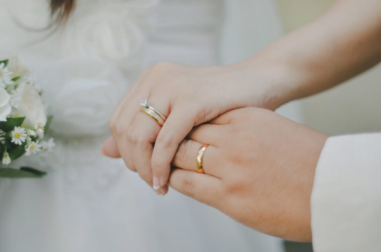 Close-up of a couple's hands with wedding bands; a bouquet of white flowers visible on the left side.