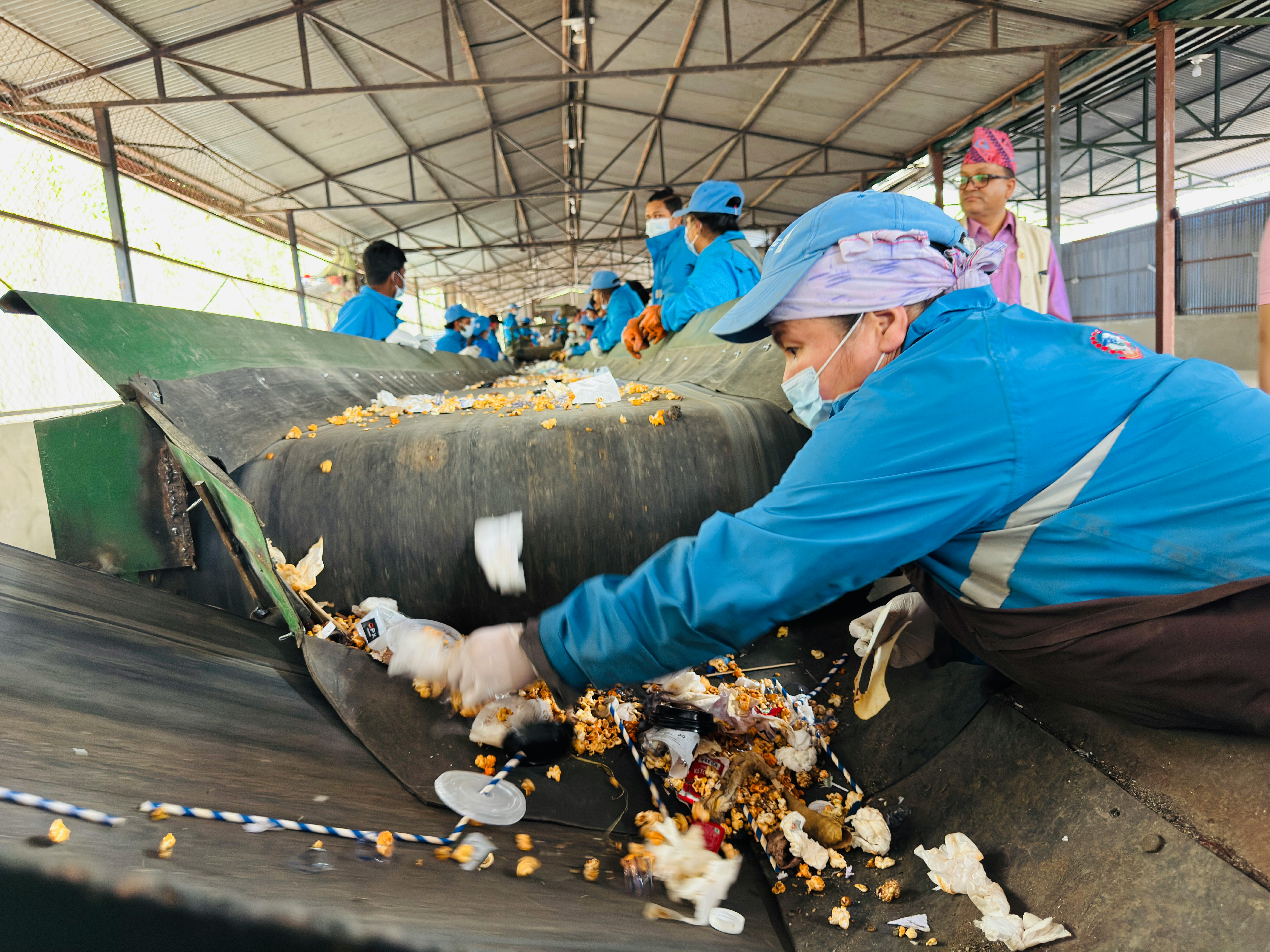 Workers in blue protective suits and masks sort trash on a moving conveyor inside a covered processing facility, picking litter from the belt.