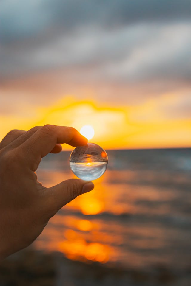Close-up of a hand holding a glass marble at sunset, reflecting the sun and ocean in the sphere.