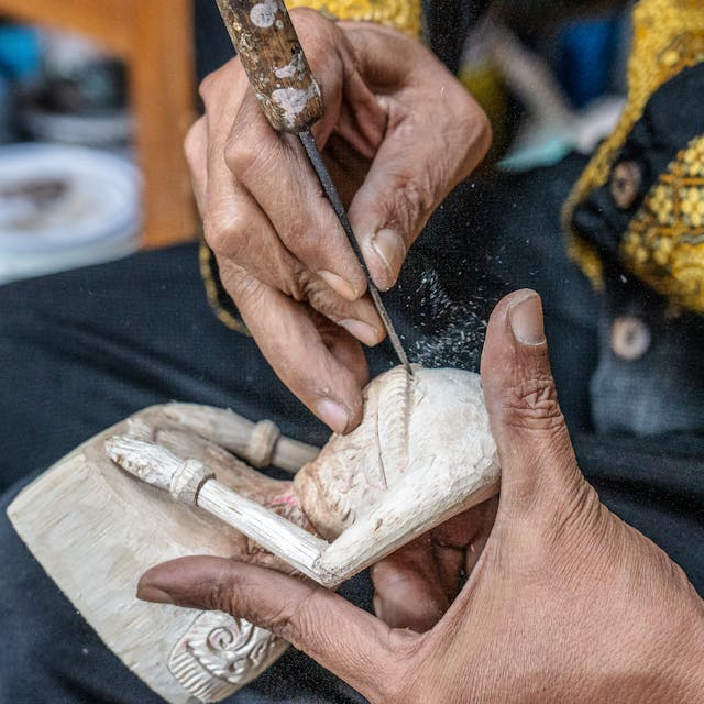 Hands carving a small wooden sculpture with a knife and chisel, focus on craftwork and toolwork detail.