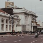 Wide city street with a white colonial-style building on the left, ornate lampposts, and busy traffic including motorcycles.