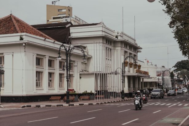Wide city street with a white colonial-style building on the left, ornate lampposts, and busy traffic including motorcycles.