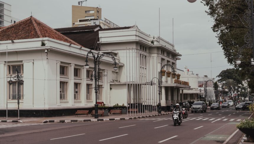 Wide city street with a white colonial-style building on the left, ornate lampposts, and busy traffic including motorcycles.