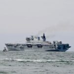 Large gray aircraft carrier sailing on calm sea with a small tugboat nearby and faint smoke from its funnel in the distance.