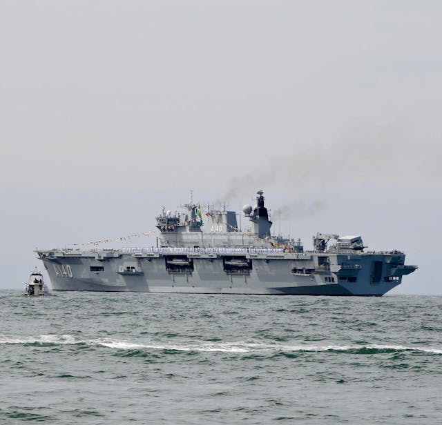 Large gray aircraft carrier sailing on calm sea with a small tugboat nearby and faint smoke from its funnel in the distance.