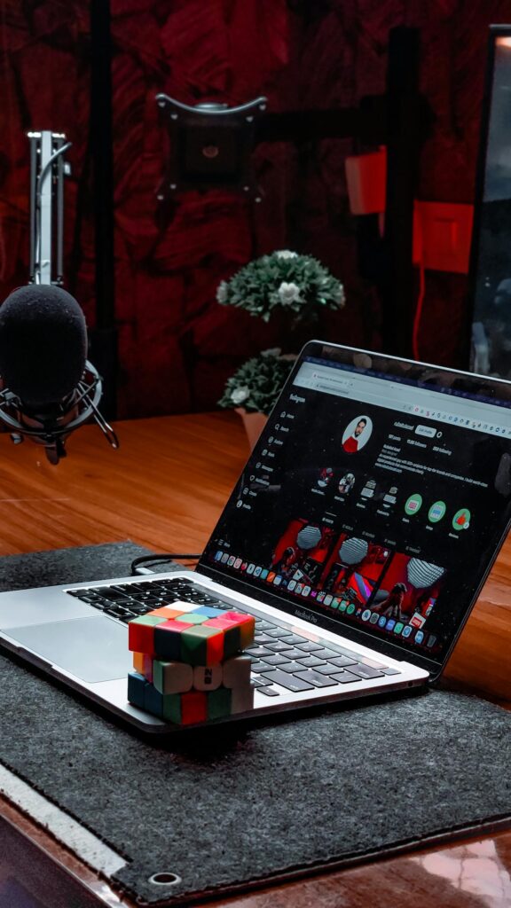 Podcast recording setup: a MacBook laptop on a desk with a colorful Rubik's cube in front, and a large microphone to the left in a red-lit studio.