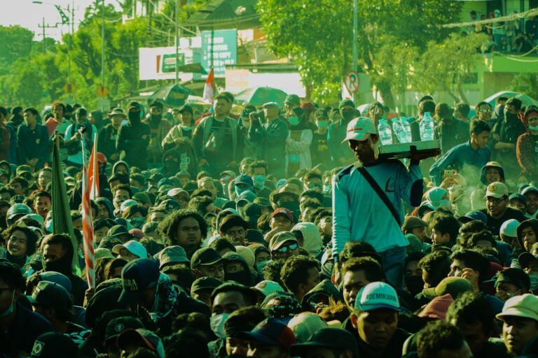 Dense crowd participating in an outdoor protest, with a man in a teal shirt and white cap carrying a water rack on his shoulder