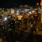 A large group of people on bicycles riding together at night on a street, smiling and looking at the camera.