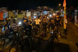 A large group of people on bicycles riding together at night on a street, smiling and looking at the camera.