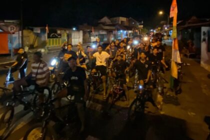 A large group of people on bicycles riding together at night on a street, smiling and looking at the camera.
