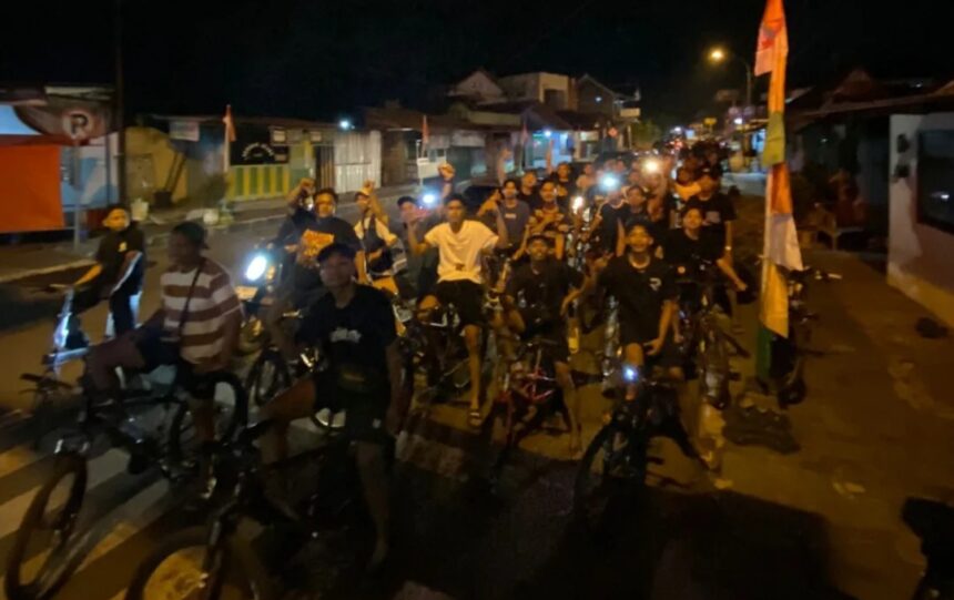 A large group of people on bicycles riding together at night on a street, smiling and looking at the camera.