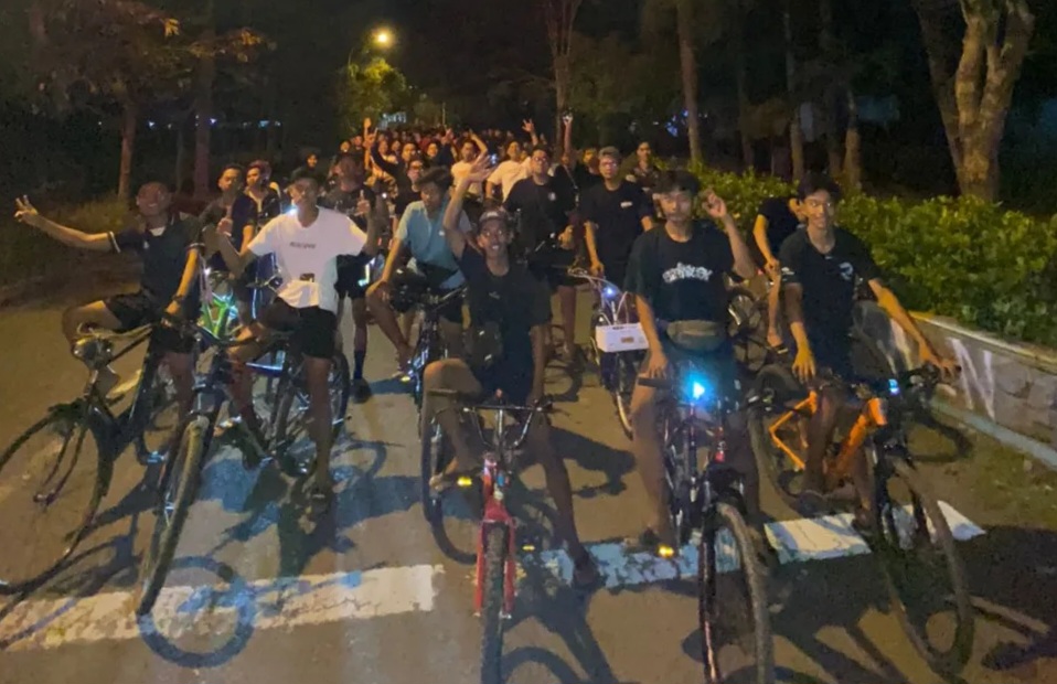 Large group of cyclists posing with their bikes on a dimly lit street at night, some waving or making peace signs.