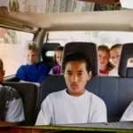 Boy in white shirt sits in the front seat of a minivan looking upset while other children lean around in the back seats.