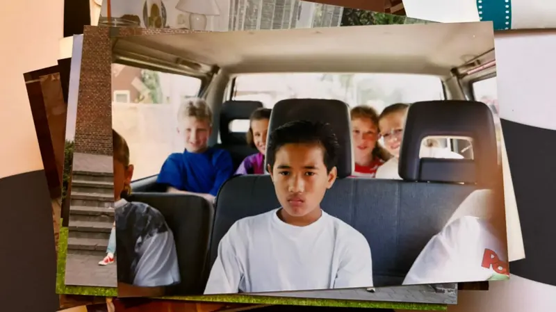 Boy in white shirt sits in the front seat of a minivan looking upset while other children lean around in the back seats.
