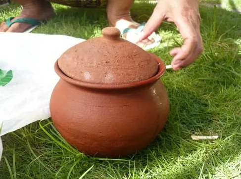 Brown clay pot with a lid sits on green grass, with hands nearby wiping or handling a cloth.