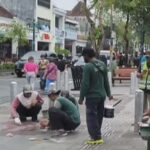 Group of workers in reflective vests repairing a sidewalk, kneeling and placing tiles while others observe with tools, across a busy street with shops and trees in the background.
