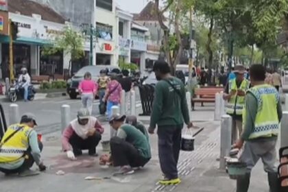 Group of workers in reflective vests repairing a sidewalk, kneeling and placing tiles while others observe with tools, across a busy street with shops and trees in the background.
