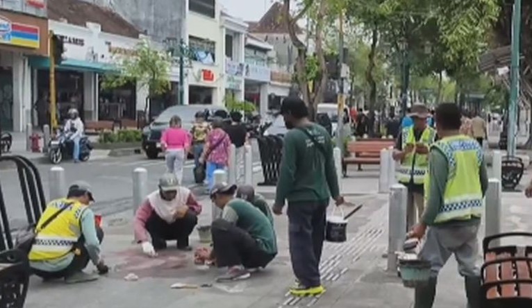 Group of workers in reflective vests repairing a sidewalk, kneeling and placing tiles while others observe with tools, across a busy street with shops and trees in the background.