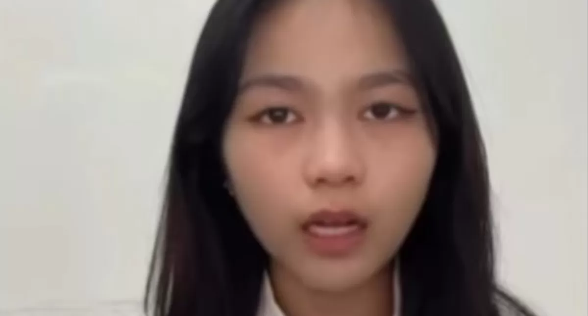 Young woman speaks directly to the camera against a light background, likely recording a video message or vlog.