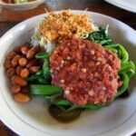 Plate of sautéed greens with a chunky peanut sauce topping, roasted peanuts, and crispy fried onions on the side.