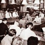 Historic mid-20th-century council meeting with men in suits seated around a long U-shaped table, many reading documents.