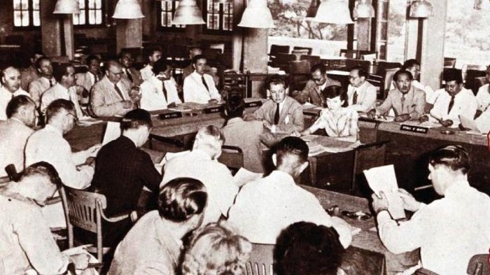 Historic mid-20th-century council meeting with men in suits seated around a long U-shaped table, many reading documents.