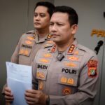 Two Indonesian police officers in beige uniforms stand with a document during a briefing, badges visible on their chests.