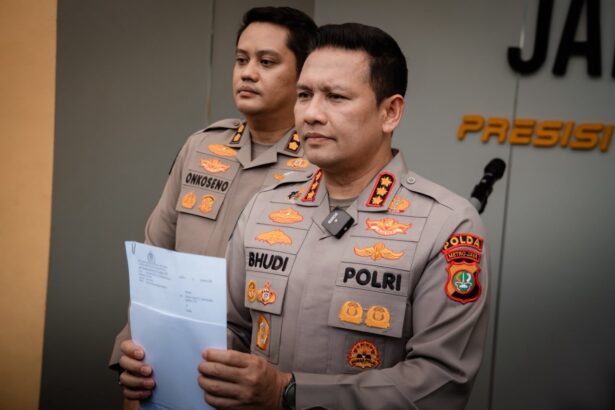 Two Indonesian police officers in beige uniforms stand with a document during a briefing, badges visible on their chests.