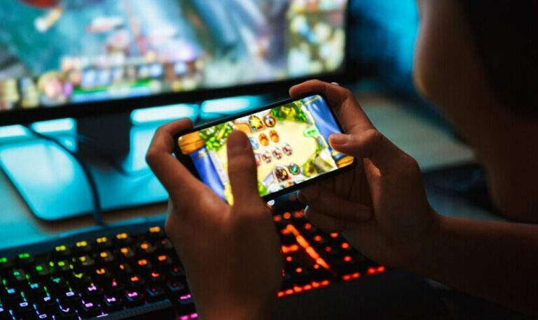 Person plays a mobile game on a smartphone, with a colorful gaming setup and RGB keyboard in the background.