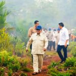 Group of officials walking along a muddy path through lush tropical plants, led by a man in a beige uniform and blue cap.