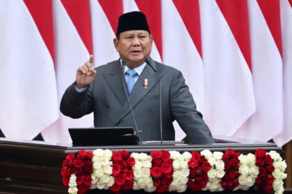 Indonesian official giving a speech at a podium decorated with red and white roses, national flags in the background.