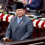 Man in a dark suit and songkok delivers a speech at a wooden podium in a formal chamber, with red and white flower garlands behind him.