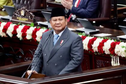 Man in a dark suit and songkok delivers a speech at a wooden podium in a formal chamber, with red and white flower garlands behind him.