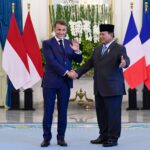 Two men in dark suits shake hands in a formal room, with red–white and blue–white–red flags behind them and a floral arrangement.