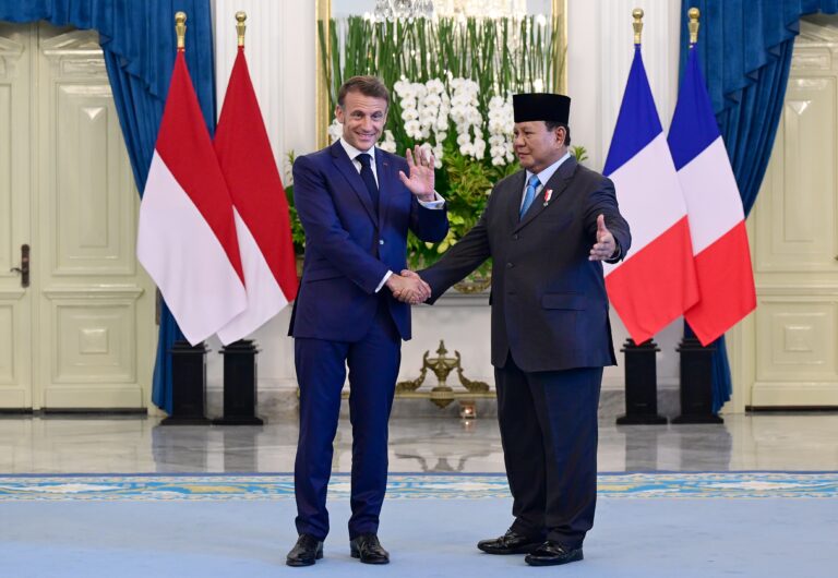 Two men in dark suits shake hands in a formal room, with red–white and blue–white–red flags behind them and a floral arrangement.