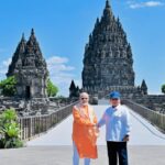 Two men in bright robes shake hands on a sunny walkway in front of tall ancient Hindu temple towers.