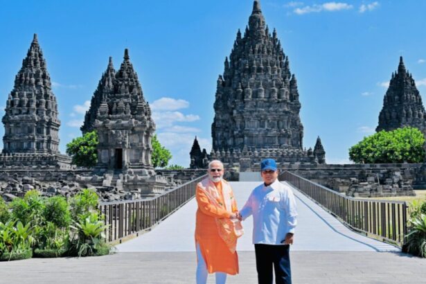 Two men in bright robes shake hands on a sunny walkway in front of tall ancient Hindu temple towers.