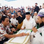 Two men shake hands across a low table as a crowd surrounds them, one holding a microphone, at a public gathering.