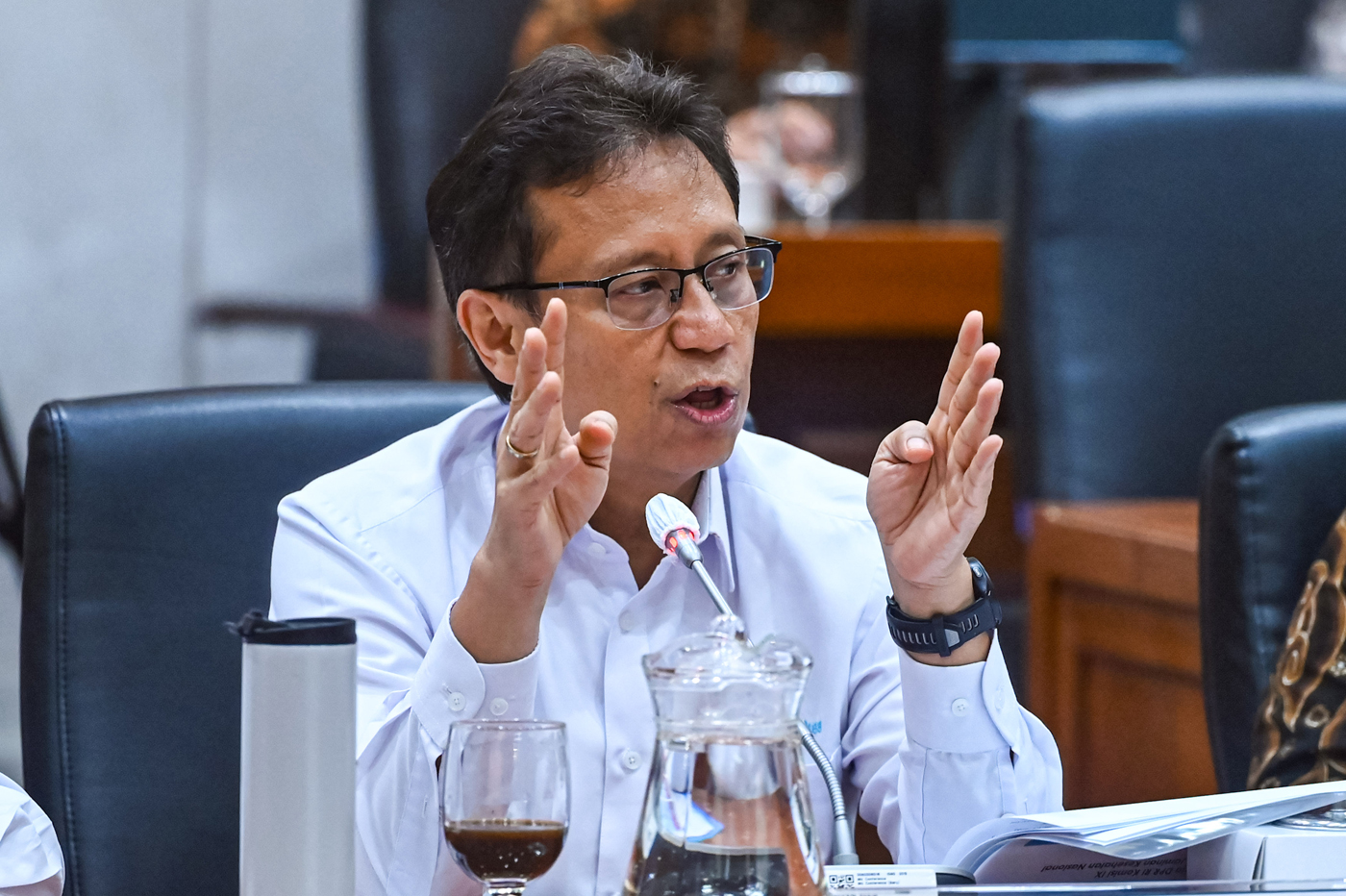 Man in glasses speaks at a meeting, hands raised as he explains, microphone in front of him