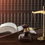 Gavel and gold scales of justice on a wooden desk with an open law book nearby.