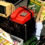 Cluttered collection of retro electronics and magazines: a beige CRT monitor, a red vintage telephone, a black appliance with a keypad, and Gun Digest magazines nearby. Also visible is a corded phone and a tray of colorful stickers in the foreground.