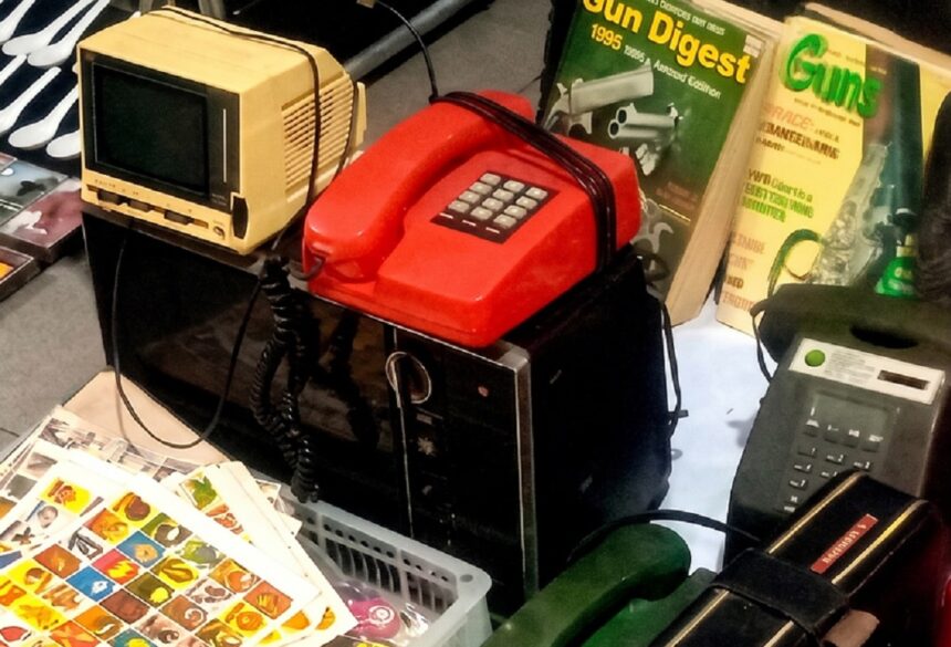 Cluttered collection of retro electronics and magazines: a beige CRT monitor, a red vintage telephone, a black appliance with a keypad, and Gun Digest magazines nearby. Also visible is a corded phone and a tray of colorful stickers in the foreground.